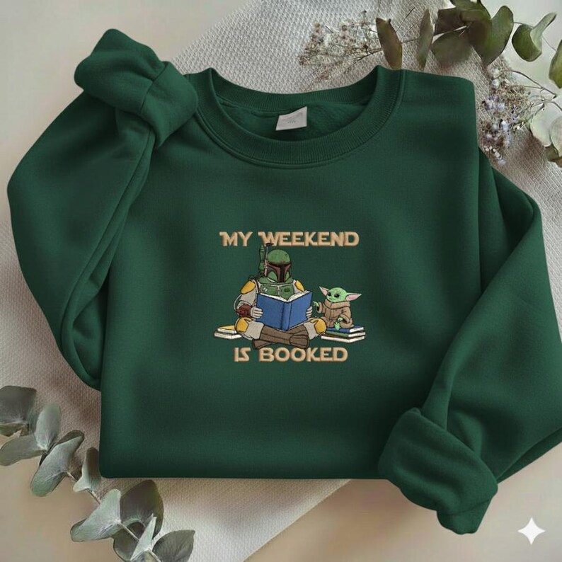 Pu&ograve; includere: Felpa verde scuro con la scritta "MY WEEKEND IS BOOKED" e un'illustrazione di un personaggio che legge un libro con una piccola creatura verde. La felpa ha maniche lunghe e girocollo.