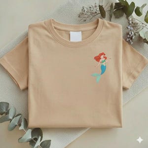 May include: A tan-colored t-shirt with a crew neck and short sleeves. The shirt features a small graphic of a mermaid with red hair, a green top, and a blue tail. The shirt is folded neatly on a light-colored surface.