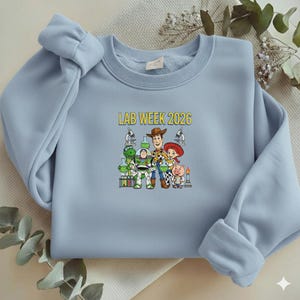 Puede incluir: Sudadera azul claro con un gr&aacute;fico de personajes de Toy Story y el texto "LAB WEEK 2026" en amarillo. El dise&ntilde;o incluye a Woody, Buzz Lightyear, Jessie y otros personajes, con equipo de laboratorio.