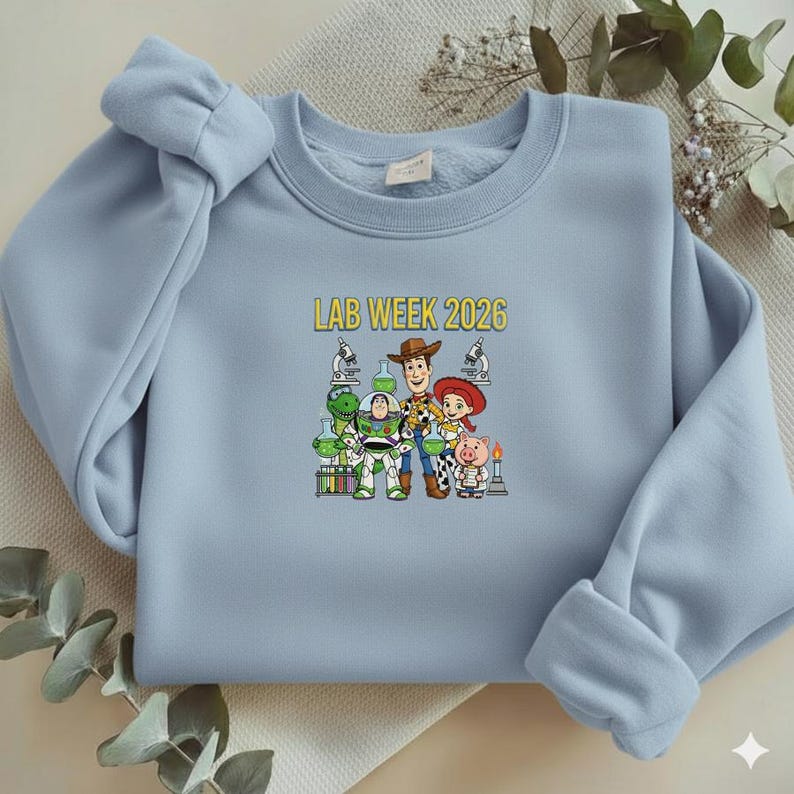 Puede incluir: Sudadera azul claro con un gr&aacute;fico de personajes de Toy Story y el texto "LAB WEEK 2026" en amarillo. El dise&ntilde;o incluye a Woody, Buzz Lightyear, Jessie y otros personajes, con equipo de laboratorio.