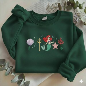 Puede incluir: Sudadera verde oscuro con un dise&ntilde;o de sirena. El dise&ntilde;o incluye una sirena con pelo rojo y cola verde, una concha, un tridente, una estrella de mar y coral. El dise&ntilde;o est&aacute; centrado en la parte delantera.