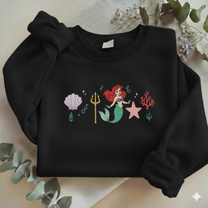 Puede incluir: Sudadera negra con un dise&ntilde;o bordado de una sirena con pelo rojo y cola verde, un tridente dorado, una estrella de mar rosa y una concha. El dise&ntilde;o tambi&eacute;n incluye burbujas y coral. La sudadera est&aacute; hecha de un material suave.