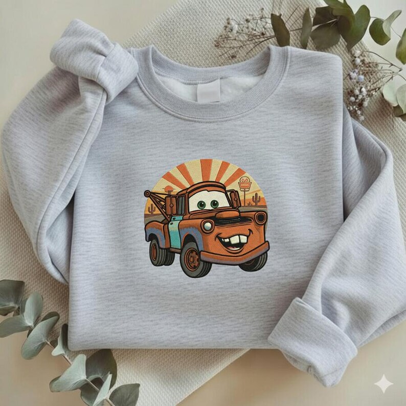 May include: Light gray sweatshirt featuring a cartoon image of Tow Mater from the Cars movie. The graphic shows Mater in orange and blue with a desert background and a rising sun design. The sweatshirt is made of a soft material.