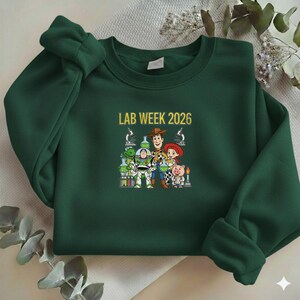 Puede incluir: Sudadera verde oscuro con el texto "LAB WEEK 2026" en dorado. El gr&aacute;fico presenta personajes de dibujos animados con equipos de laboratorio. Los personajes incluyen un dinosaurio verde, Buzz Lightyear, Woody, Jessie y un cerdo.