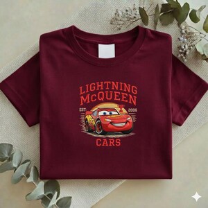Puede incluir: Una camiseta granate con un dise&ntilde;o de Rayo McQueen de la pel&iacute;cula Cars. El dise&ntilde;o incluye el texto "LIGHTNING McQUEEN" encima de la imagen del coche, con "EST. 2006" y "CARS" debajo. La camiseta est&aacute; colocada sobre una superficie neutra.