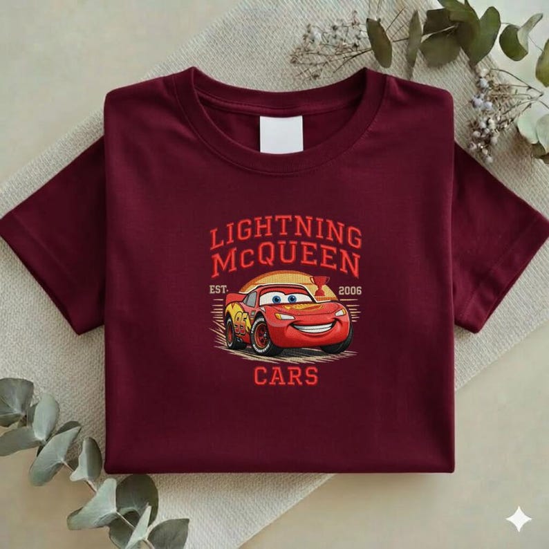 Puede incluir: Una camiseta granate con un dise&ntilde;o de Rayo McQueen de la pel&iacute;cula Cars. El dise&ntilde;o incluye el texto "LIGHTNING McQUEEN" encima de la imagen del coche, con "EST. 2006" y "CARS" debajo. La camiseta est&aacute; colocada sobre una superficie neutra.