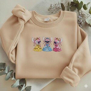 Puede incluir: Sudadera beige con un dise&ntilde;o bordado de tres personajes de dibujos animados con vestidos de princesa. Los personajes son rosas y azules con coronas y vestidos amarillos, azules y rosas. La sudadera tiene cuello redondo y mangas largas.