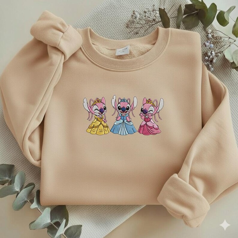 Puede incluir: Sudadera beige con un dise&ntilde;o bordado de tres personajes de dibujos animados con vestidos de princesa. Los personajes son rosas y azules con coronas y vestidos amarillos, azules y rosas. La sudadera tiene cuello redondo y mangas largas.