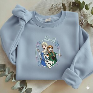 Puede incluir: Sudadera azul claro con un gr&aacute;fico circular de los personajes animados Elsa, Anna y Olaf. El gr&aacute;fico est&aacute; rodeado por una corona floral con un lazo en la parte superior. La sudadera tiene mangas largas y cuello redondo.