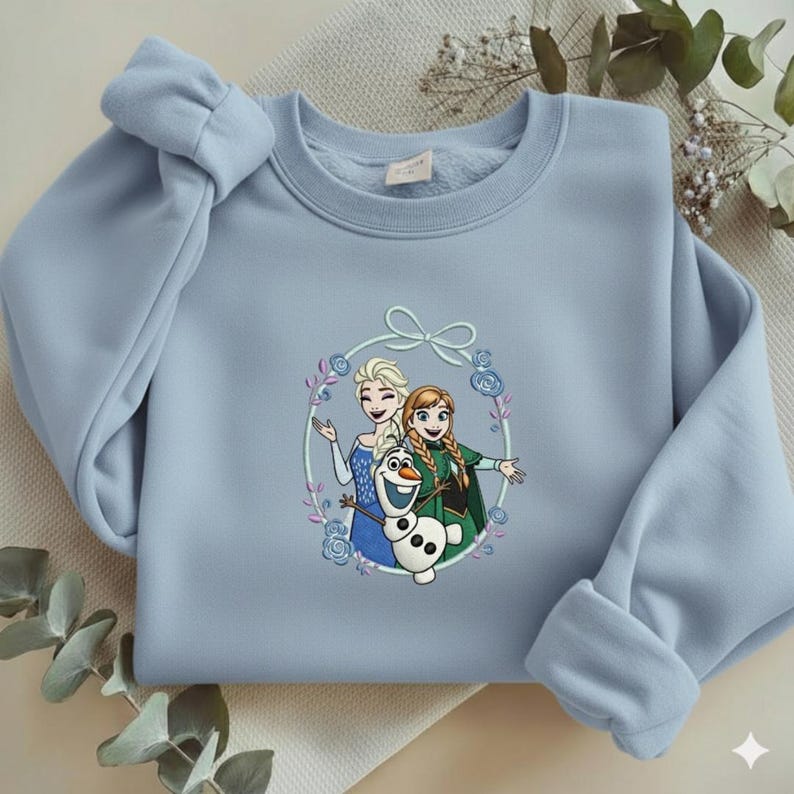Puede incluir: Sudadera azul claro con un gr&aacute;fico circular de los personajes animados Elsa, Anna y Olaf. El gr&aacute;fico est&aacute; rodeado por una corona floral con un lazo en la parte superior. La sudadera tiene mangas largas y cuello redondo.