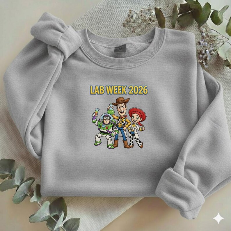May include: A light gray sweatshirt featuring a cartoon graphic of Buzz Lightyear, Woody, and Jessie. The text "LAB WEEK 2026" is printed above the characters in yellow. The sweatshirt has long sleeves and a crew neck.