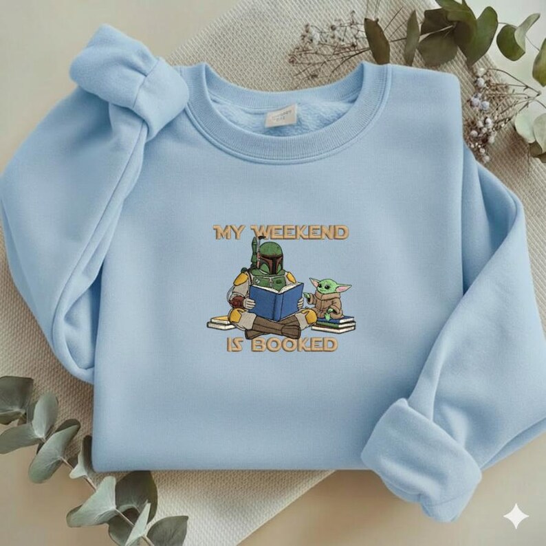 Pu&ograve; includere: Felpa azzurra con la scritta "MY WEEKEND IS BOOKED". La grafica mostra un personaggio dei cartoni animati con un'armatura verde che legge un libro con una piccola creatura verde. La felpa ha un girocollo e maniche lunghe.