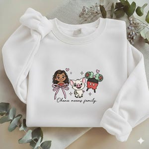 Puede incluir: Sudadera blanca con personajes de dibujos animados bordados y la frase "Ohana means family". El dise&ntilde;o incluye una ni&ntilde;a con un lazo rosa, un cerdo y un personaje con un lazo. La sudadera tiene mangas largas y cuello redondo.