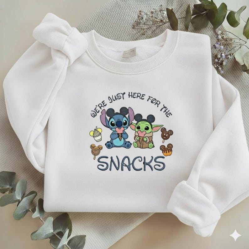 May include: White sweatshirt featuring a graphic of Stitch and Grogu with the text "WE'RE JUST HERE FOR THE SNACKS." The characters are holding ice cream and snacks. The sweatshirt has long sleeves and a crew neck.