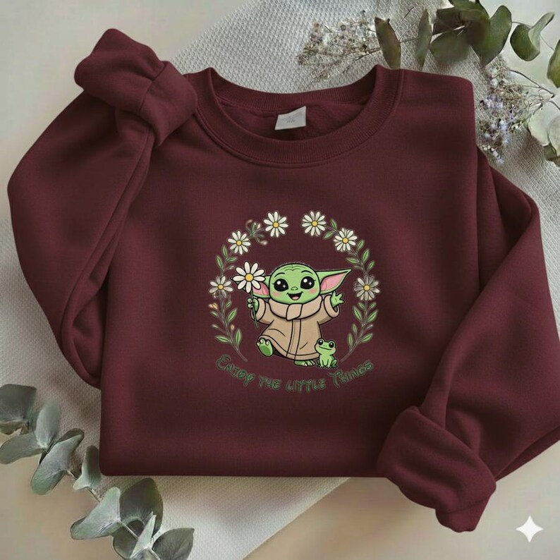 May include: Burgundy sweatshirt featuring a graphic of a green alien character surrounded by a daisy wreath, holding a flower and a small frog. The text "Enjoy the little things" is below the graphic.