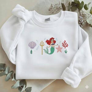 Puede incluir: Sudadera blanca con un dise&ntilde;o bordado. El dise&ntilde;o presenta una sirena con cabello rojo y cola verde, un tridente dorado, una estrella de mar rosa y coral. Tambi&eacute;n incluye una concha y burbujas.