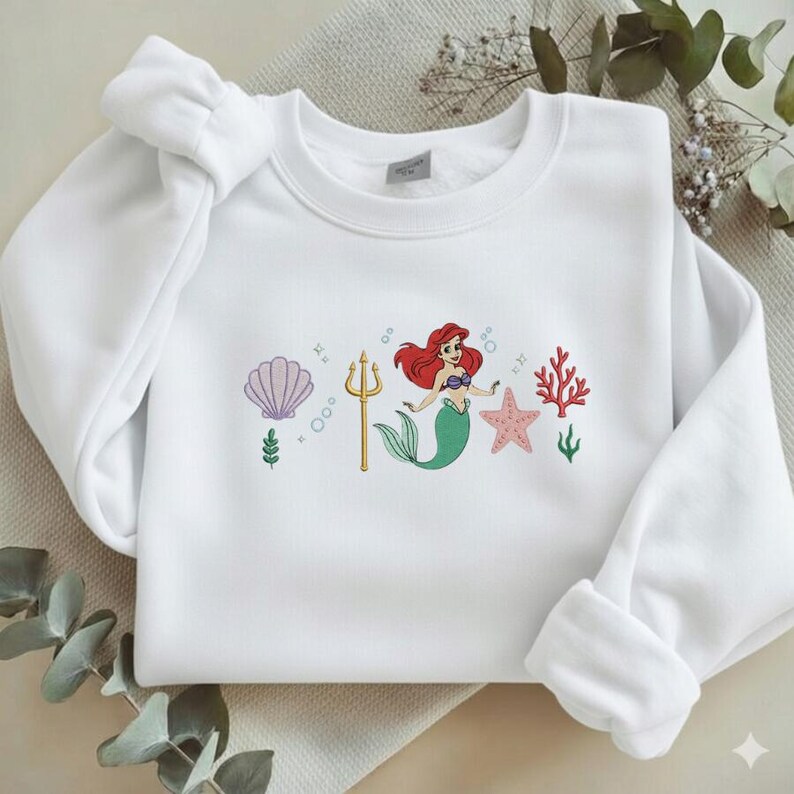 Puede incluir: Sudadera blanca con un dise&ntilde;o bordado. El dise&ntilde;o presenta una sirena con cabello rojo y cola verde, un tridente dorado, una estrella de mar rosa y coral. Tambi&eacute;n incluye una concha y burbujas.