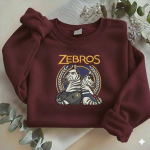 Może przedstawiać: Bordowa bluza z napisem "ZEBROS" w złotym kolorze nad grafiką dw&oacute;ch zebr w mundurach. Bluza ma długie rękawy i okrągły dekolt. Projekt jest wyśrodkowany.