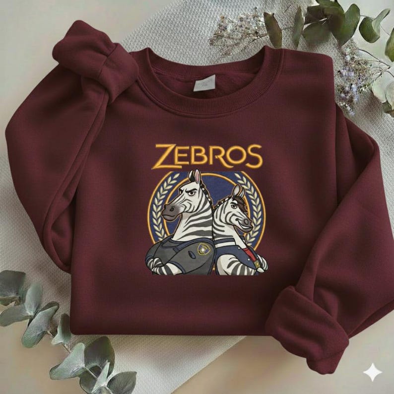 Może przedstawiać: Bordowa bluza z napisem "ZEBROS" w złotym kolorze nad grafiką dw&oacute;ch zebr w mundurach. Bluza ma długie rękawy i okrągły dekolt. Projekt jest wyśrodkowany.
