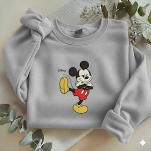 Puede incluir: Sudadera gris claro con un bordado de Mickey Mouse. Mickey est&aacute; representado en su pose cl&aacute;sica, con pantalones cortos rojos y zapatos amarillos. El logotipo de Disney est&aacute; impreso en la parte superior izquierda.