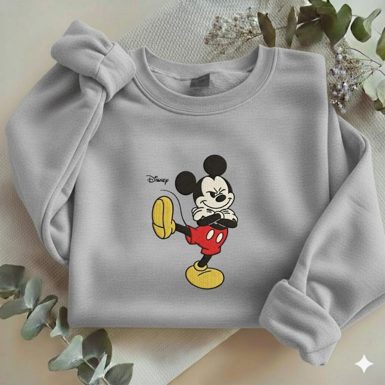Puede incluir: Sudadera gris claro con un bordado de Mickey Mouse. Mickey est&aacute; representado en su pose cl&aacute;sica, con pantalones cortos rojos y zapatos amarillos. El logotipo de Disney est&aacute; impreso en la parte superior izquierda.