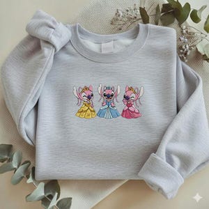 Puede incluir: Una sudadera gris claro con un dise&ntilde;o bordado de tres personajes de dibujos animados con vestidos de princesa. Los vestidos son amarillos, azules y rosas. Los personajes tienen piel rosa y llevan coronas.
