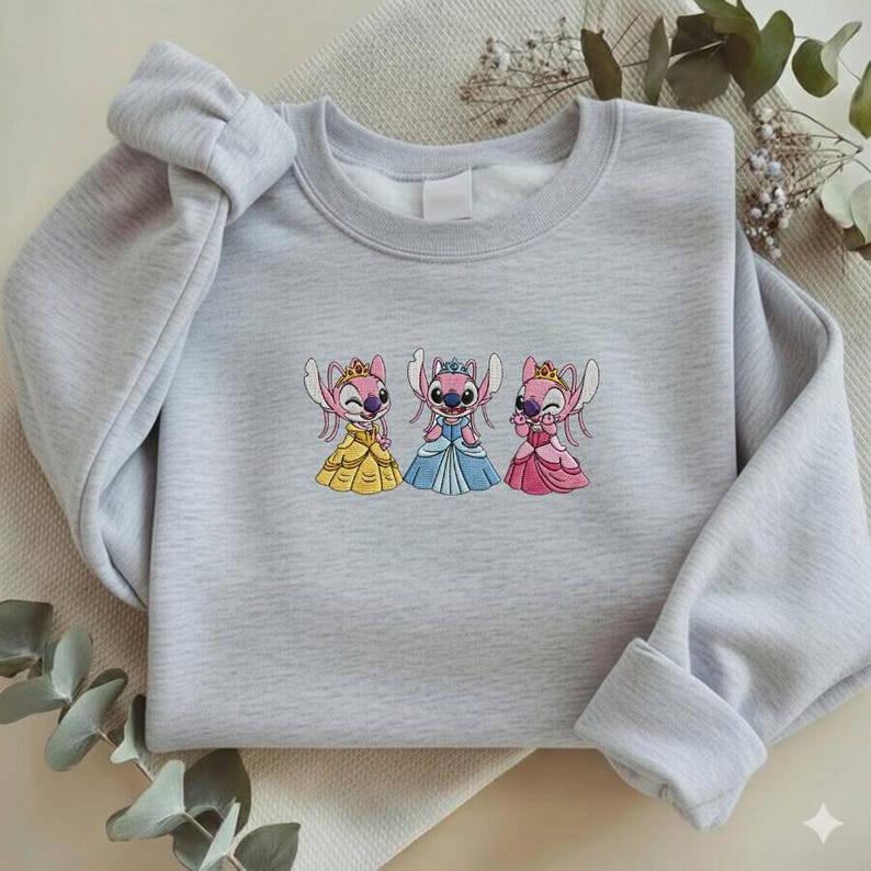 Puede incluir: Una sudadera gris claro con un dise&ntilde;o bordado de tres personajes de dibujos animados con vestidos de princesa. Los vestidos son amarillos, azules y rosas. Los personajes tienen piel rosa y llevan coronas.