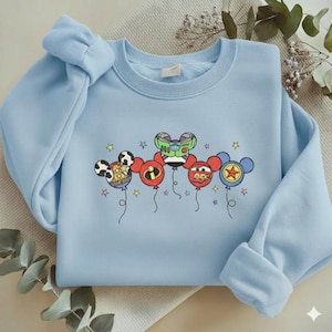 Może przedstawiać: Jasnoniebieska bluza z balonami z postaciami z kresk&oacute;wek. Balony zaprojektowano jako Myszka Miki, Buzz Lightyear i Zygzak McQueen. Bluza ma okrągły dekolt i długie rękawy.