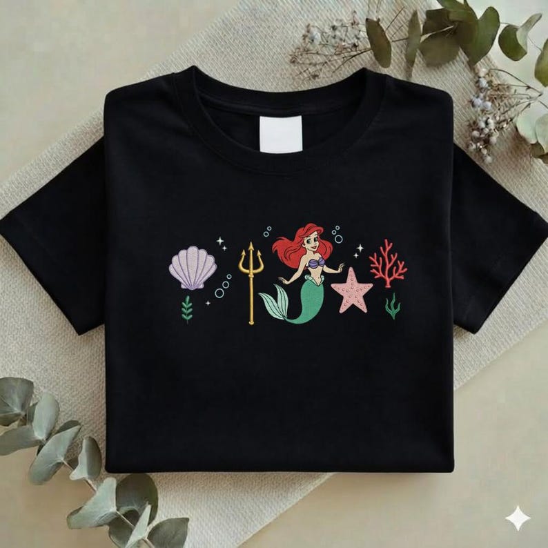 Puede incluir: Camiseta negra con un gr&aacute;fico colorido de una sirena con cabello rojo y cola verde, junto con una concha, un tridente, una estrella de mar y coral. El dise&ntilde;o es una ilustraci&oacute;n caprichosa.