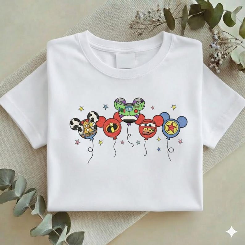 Może przedstawiać: Biała koszulka z kolorową grafiką balon&oacute;w z postaciami z kresk&oacute;wek. Balony, zaprojektowane z uszami Myszki Miki, reprezentują postacie takie jak Buzz Lightyear i Zygzak McQueen. Małe gwiazdki są rozrzucone wok&oacute;ł balon&oacute;w.