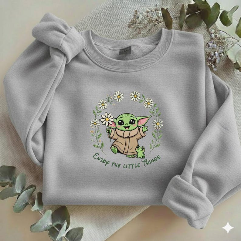 May include: A light gray sweatshirt featuring a cartoon character holding a daisy, surrounded by a floral wreath. The text "Enjoy the little things" is printed below the character. The sweatshirt has long sleeves.