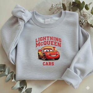 Puede incluir: Una sudadera gris claro con el texto "LIGHTNING McQUEEN" sobre una imagen de dibujos animados de un coche de carreras rojo. La palabra "CARS" est&aacute; impresa debajo. La sudadera tiene mangas largas.