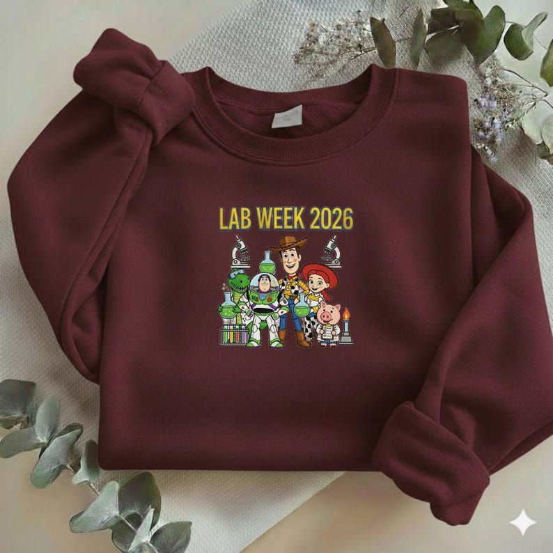 Puede incluir: Una sudadera granate con el texto "LAB WEEK 2026" en dorado. El gr&aacute;fico presenta personajes de dibujos animados con equipos de laboratorio. La sudadera est&aacute; hecha de un material suave.