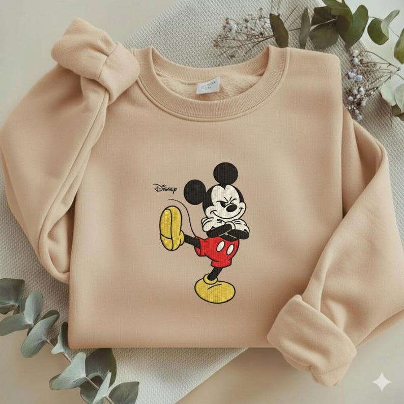 Puede incluir: Sudadera beige con un dise&ntilde;o bordado de Mickey Mouse. El logotipo de Disney est&aacute; impreso en la parte superior izquierda. La sudadera tiene cuello redondo y mangas largas con un detalle de lazo en el hombro izquierdo. Mickey lleva pantalones cortos rojos.