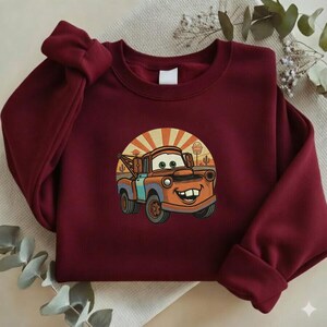May include: A maroon sweatshirt featuring a cartoon image of Mater from the Cars movie. The graphic shows Mater with a smiling face, set against a desert landscape with a rising sun design. The sweatshirt has a crew neck and long sleeves.