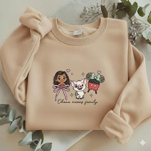 Puede incluir: Sudadera beige con personajes de dibujos animados bordados y la frase "Ohana means family". El dise&ntilde;o incluye una ni&ntilde;a con un lazo rosa, un cerdo y un personaje inspirado en Mickey Mouse con un lazo. La sudadera tiene cuello redondo y mangas largas.