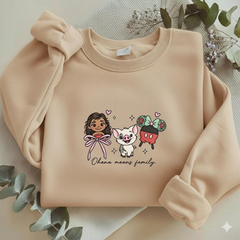 Puede incluir: Sudadera beige con personajes de dibujos animados bordados y la frase "Ohana means family". El dise&ntilde;o incluye una ni&ntilde;a con un lazo rosa, un cerdo y un personaje inspirado en Mickey Mouse con un lazo. La sudadera tiene cuello redondo y mangas largas.