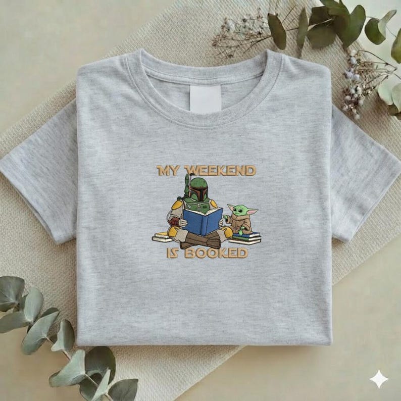 Pu&ograve; includere: T-shirt grigio chiaro con una grafica di Boba Fett e Grogu che leggono un libro. Il testo "MY WEEKEND IS BOOKED" &egrave; sopra e sotto l'illustrazione. La maglietta &egrave; piegata.