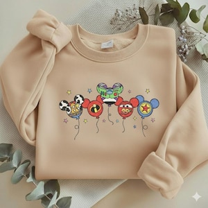 Może przedstawiać: Beżowa bluza z kolorową grafiką balon&oacute;w z postaciami z kresk&oacute;wek. Balony mają twarze postaci i gwiazdki. Bluza ma okrągły dekolt, długie rękawy i kokardę na ramieniu.