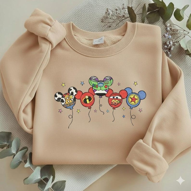 Może przedstawiać: Beżowa bluza z kolorową grafiką balon&oacute;w z postaciami z kresk&oacute;wek. Balony mają twarze postaci i gwiazdki. Bluza ma okrągły dekolt, długie rękawy i kokardę na ramieniu.