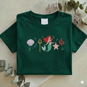 Puede incluir: Camiseta verde oscuro con un dise&ntilde;o de una sirena pelirroja, una concha, un tridente, una estrella de mar y coral. El dise&ntilde;o est&aacute; centrado en la parte delantera de la camiseta.