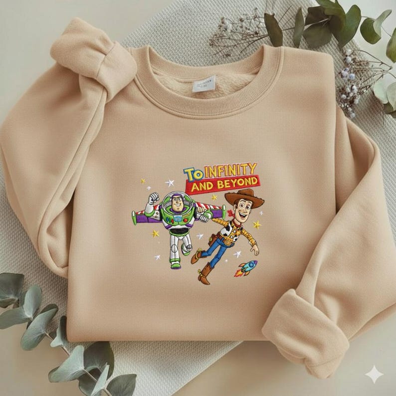 K&ouml;nnte beinhalten: Beiges Sweatshirt mit einem farbenfrohen Aufdruck von Buzz Lightyear und Woody aus Toy Story, mit dem Text "TO INFINITY AND BEYOND". Das Sweatshirt hat einen Rundhalsausschnitt und lange &Auml;rmel mit gerollten B&uuml;ndchen.