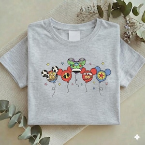 Może przedstawiać: Jasnoszara koszulka z kolorową grafiką balon&oacute;w z postaciami z kresk&oacute;wek. Balony zawierają wzory Myszki Miki, Buzz Lightyear i Zygzaka McQueena. Koszulka jest złożona i leży płasko na neutralnej powierzchni.