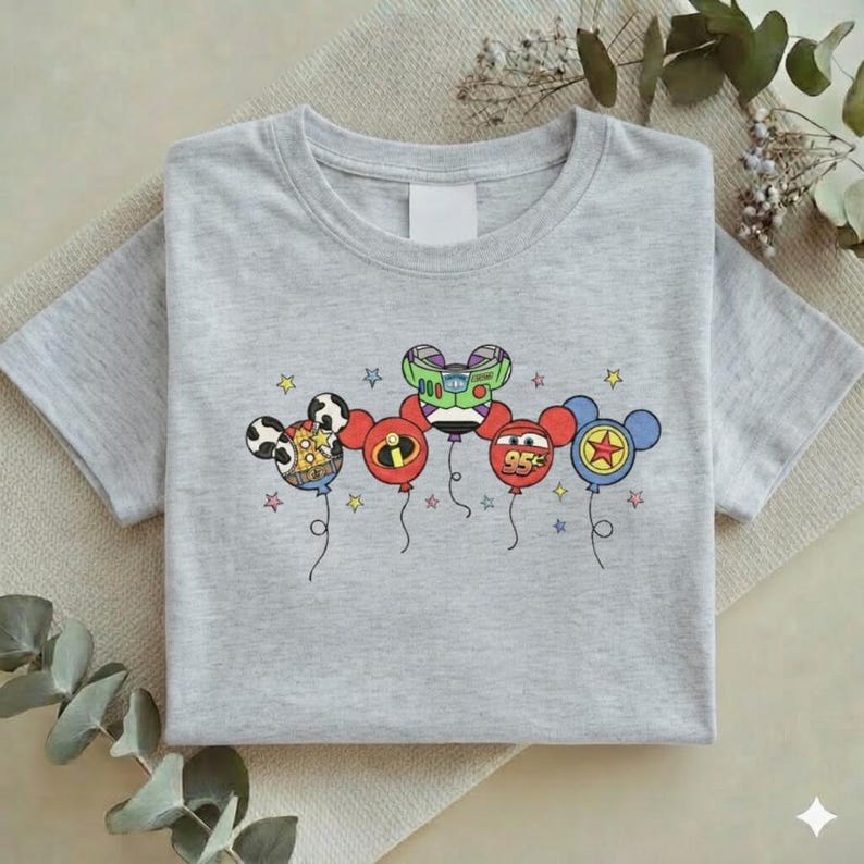 Może przedstawiać: Jasnoszara koszulka z kolorową grafiką balon&oacute;w z postaciami z kresk&oacute;wek. Balony zawierają wzory Myszki Miki, Buzz Lightyear i Zygzaka McQueena. Koszulka jest złożona i leży płasko na neutralnej powierzchni.