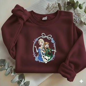 Puede incluir: Una sudadera granate con un gr&aacute;fico circular de Elsa, Anna y Olaf de la pel&iacute;cula Frozen. El gr&aacute;fico est&aacute; rodeado por una corona floral y un lazo. La sudadera tiene mangas largas.