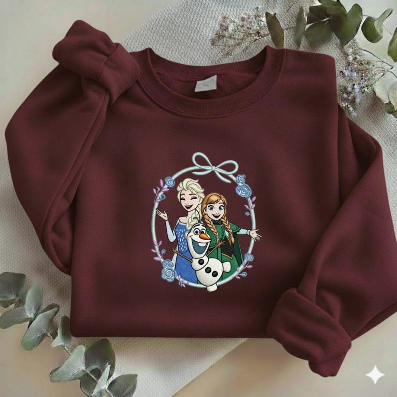 Puede incluir: Una sudadera granate con un gr&aacute;fico circular de Elsa, Anna y Olaf de la pel&iacute;cula Frozen. El gr&aacute;fico est&aacute; rodeado por una corona floral y un lazo. La sudadera tiene mangas largas.