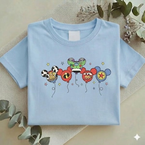 Może przedstawiać: Jasnoniebieska koszulka z grafiką balon&oacute;w z postaciami z kresk&oacute;wek. Balony zaprojektowano jako głowy Myszki Miki, z kt&oacute;rych każda reprezentuje inną postać. Projekt zawiera gwiazdy i szczeg&oacute;ły postaci.