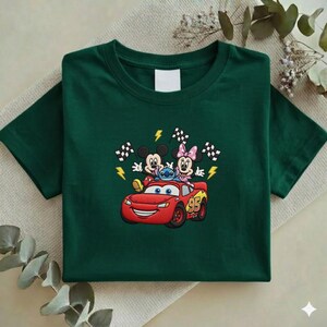 May include: Dark green t-shirt featuring an embroidered design of Mickey Mouse, Minnie Mouse, and Stitch riding in a red Lightning McQueen race car. The design includes checkered flags and lightning bolts.