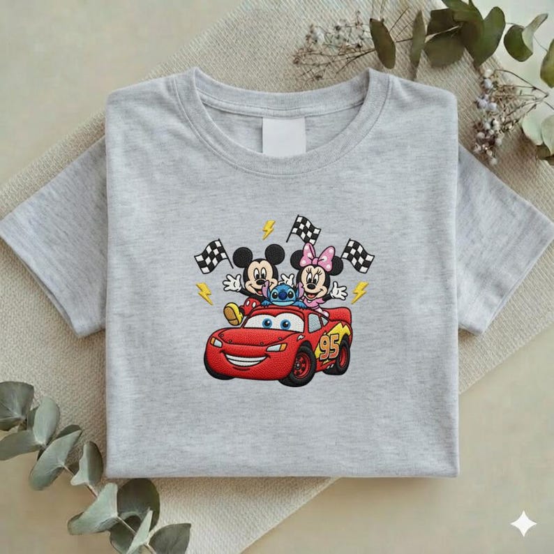 May include: A light gray t-shirt featuring an embroidered design of Mickey Mouse, Minnie Mouse, and Stitch riding in a red race car. The car has the number 95 on the side. Checkered flags and lightning bolts are also included in the design.