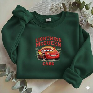 Puede incluir: Una sudadera verde oscuro con un dise&ntilde;o de Rayo McQueen de la pel&iacute;cula Cars. El dise&ntilde;o incluye el texto "LIGHTNING McQUEEN" en rojo, con "EST. 2006" y "CARS" debajo de la imagen del coche.